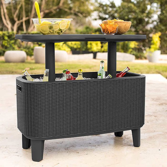 A black, woven cooler holds drinks on ice beneath a table. Above, a bowl with salad and a tray of snacks rests in an outdoor setting.