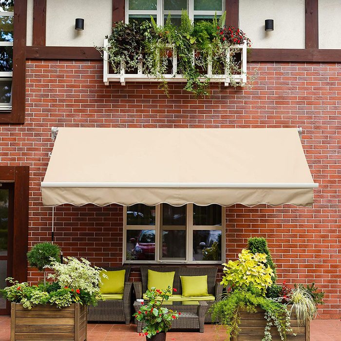 A beige awning extends over a patio seating area with green cushions, surrounded by potted plants against a red brick wall and large windows.