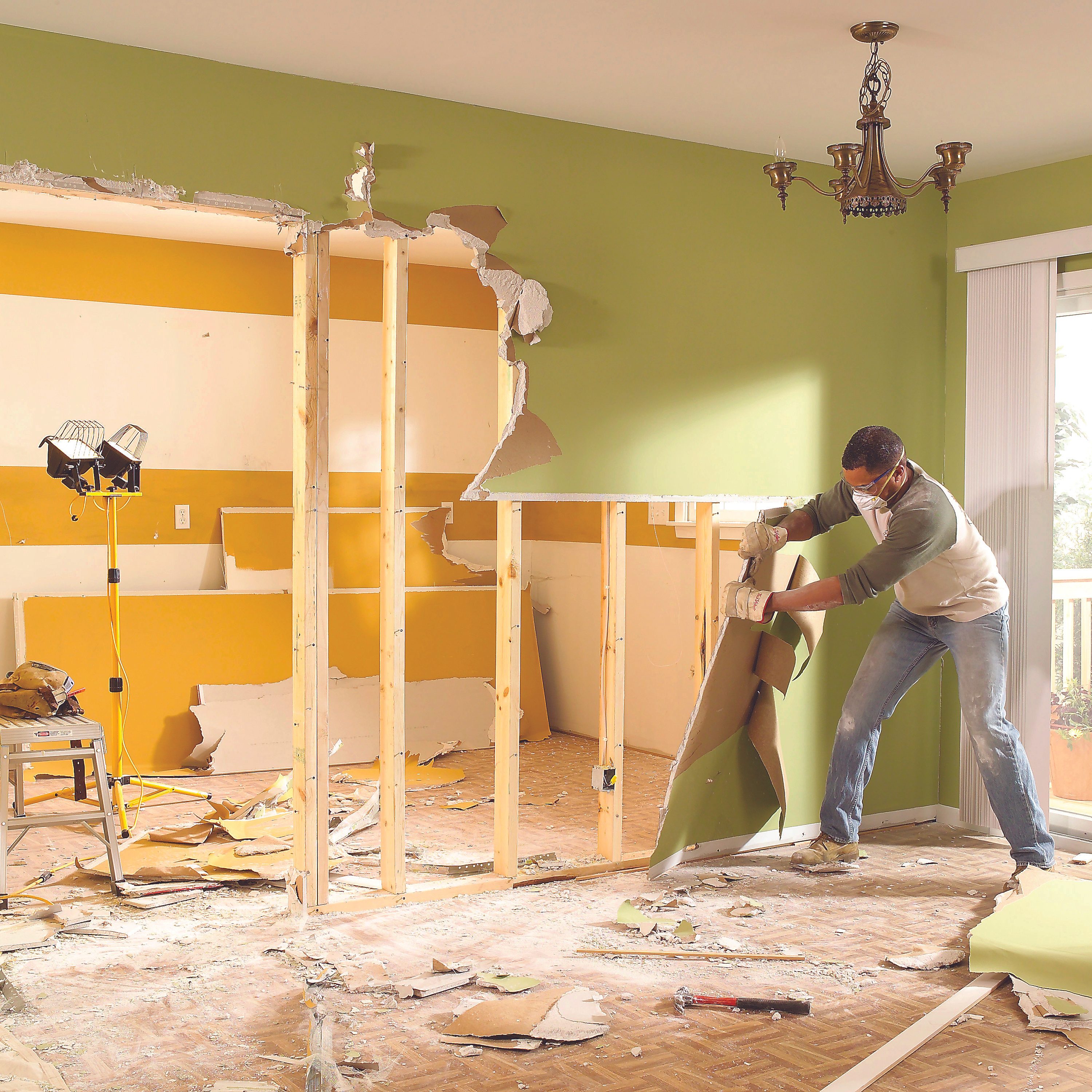A man wearing gloves removes drywall in a partially demolished room, surrounded by debris and construction tools, under a chandelier and natural light from a window.
