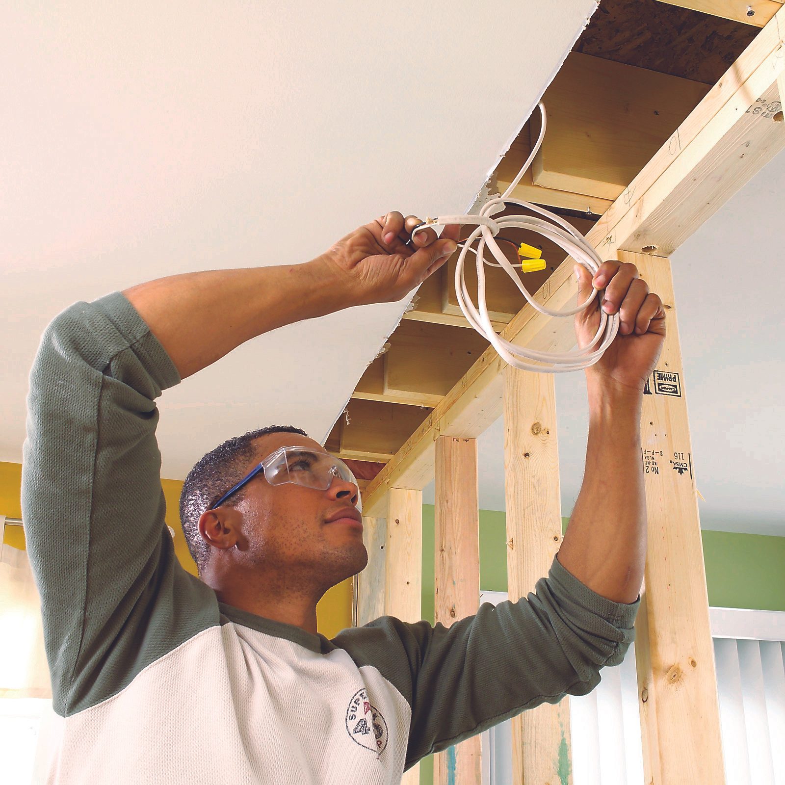 A person wearing safety glasses attaches wires to the ceiling frame inside a room, surrounded by wooden beams and green walls.