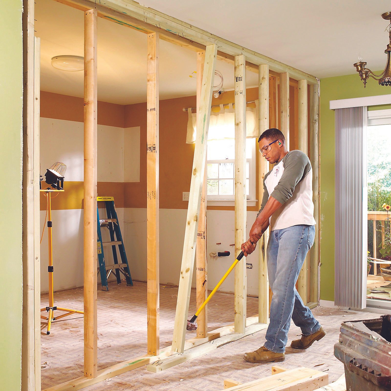 A man uses a pry bar to remove wooden framing in a partially constructed room, surrounded by tools, a ladder, and natural light from a window.