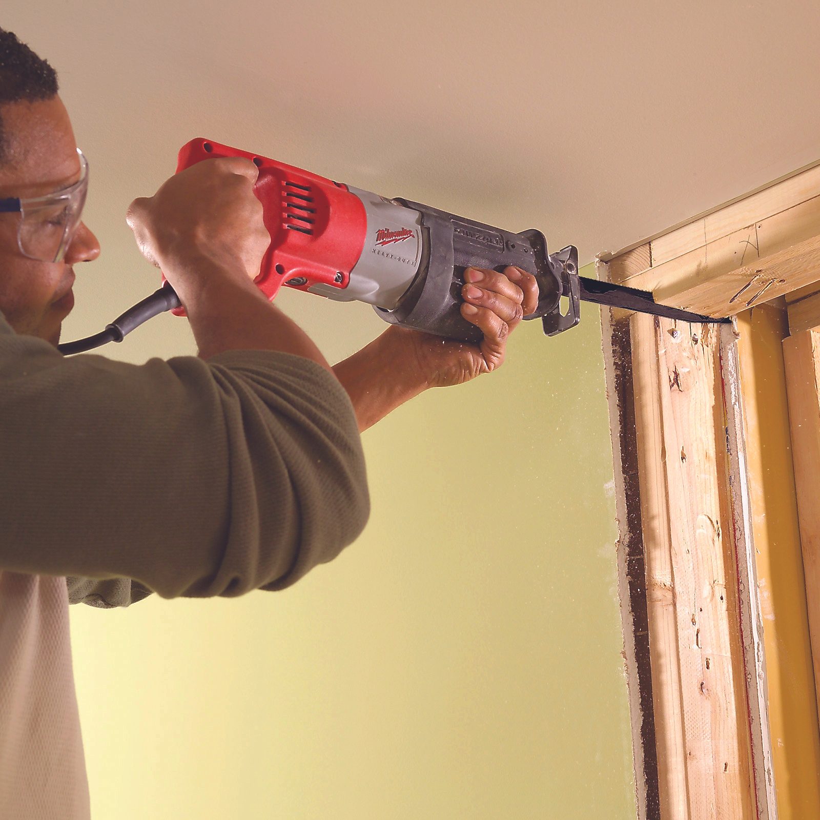 A person operates a reciprocating saw, cutting through wood near a doorway, surrounded by a green wall and wooden framing, with dust particles visible in the air.