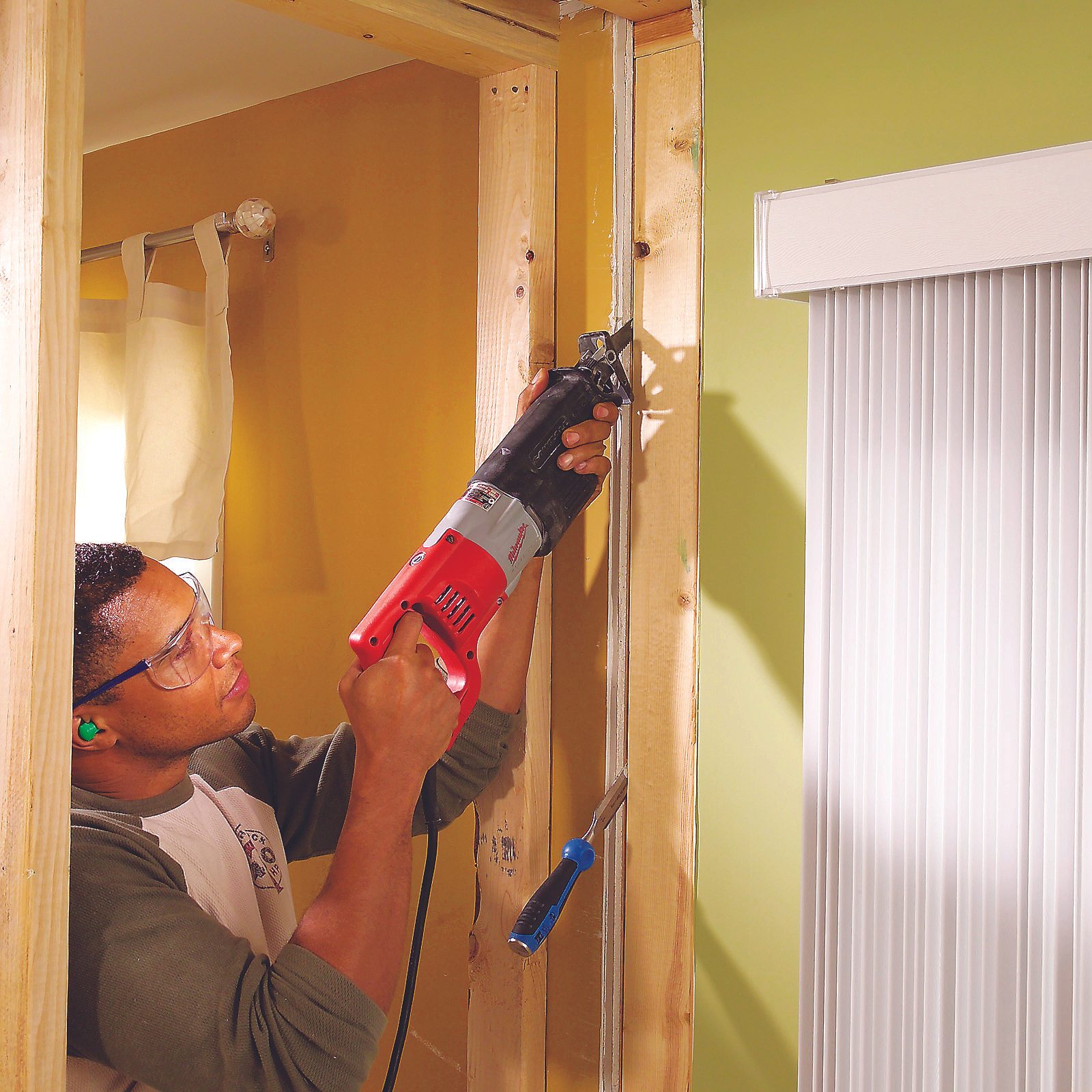 A person operates a power saw, cutting through wooden framing in a room with yellow walls and light filtering through a window with blinds.