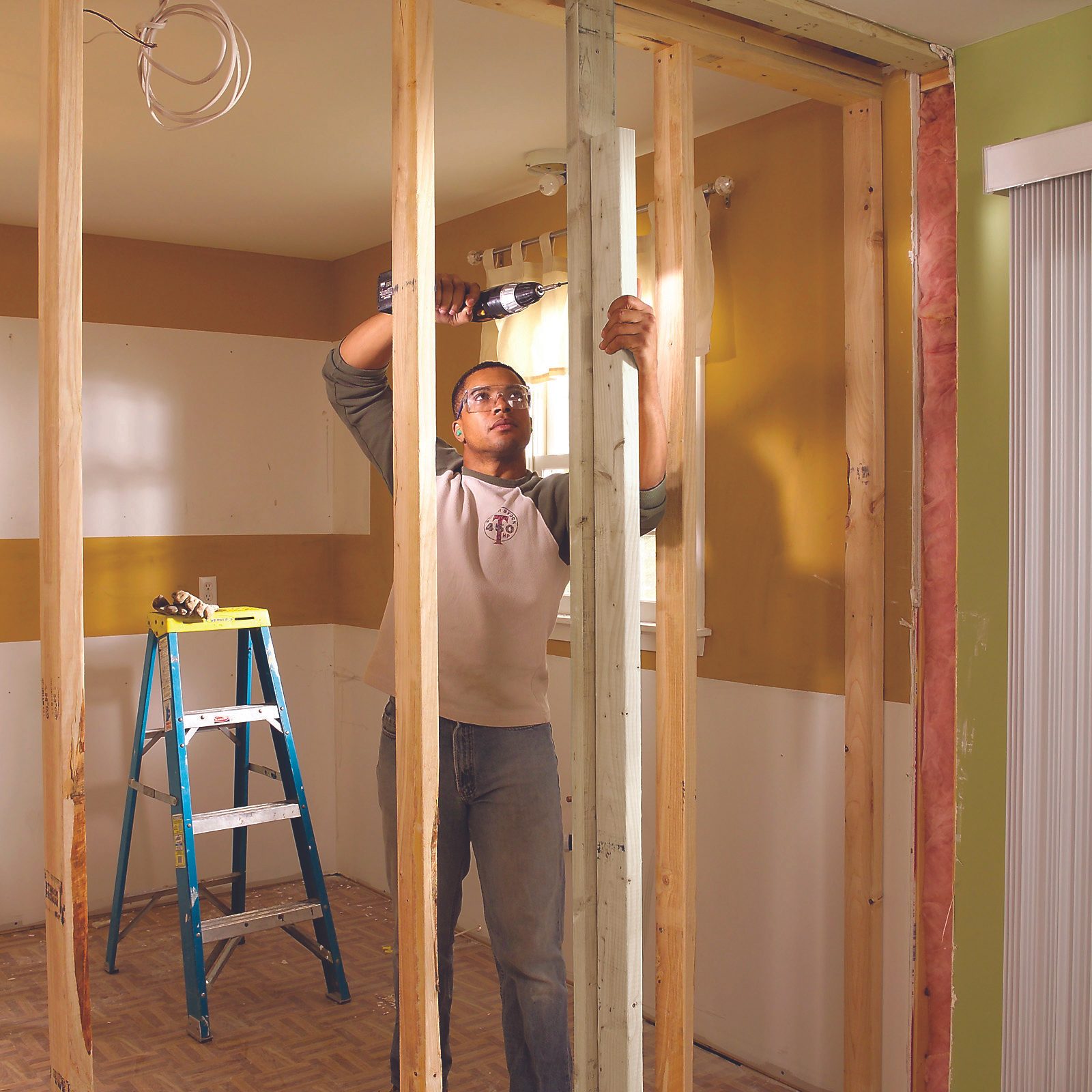 A man drills a wooden beam inside a framed room, surrounded by unfinished walls and a ladder. Natural light enters through a window nearby.