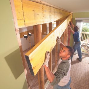 Two men lift a wooden beam above a partition in a home. Construction tools and debris are visible in a partially renovated interior space.