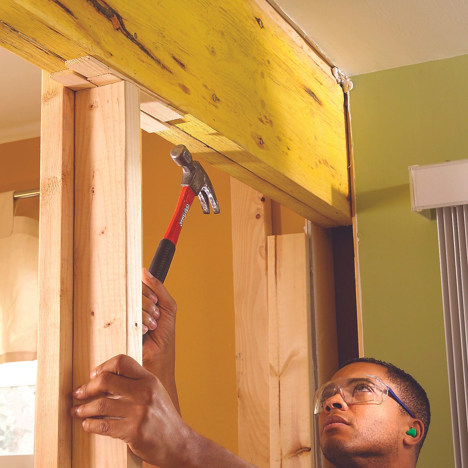 A person is hammering a wooden beam into place within a wall framework in a room with yellow and green walls.