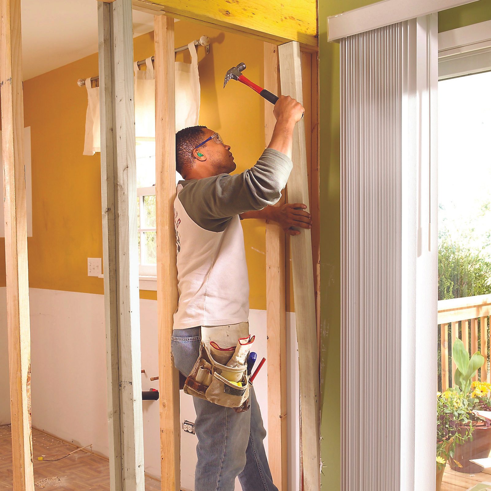 A man hammers a wooden plank in a partially renovated room with yellow walls, featuring a nearby window and a sliding door to an outdoor deck.