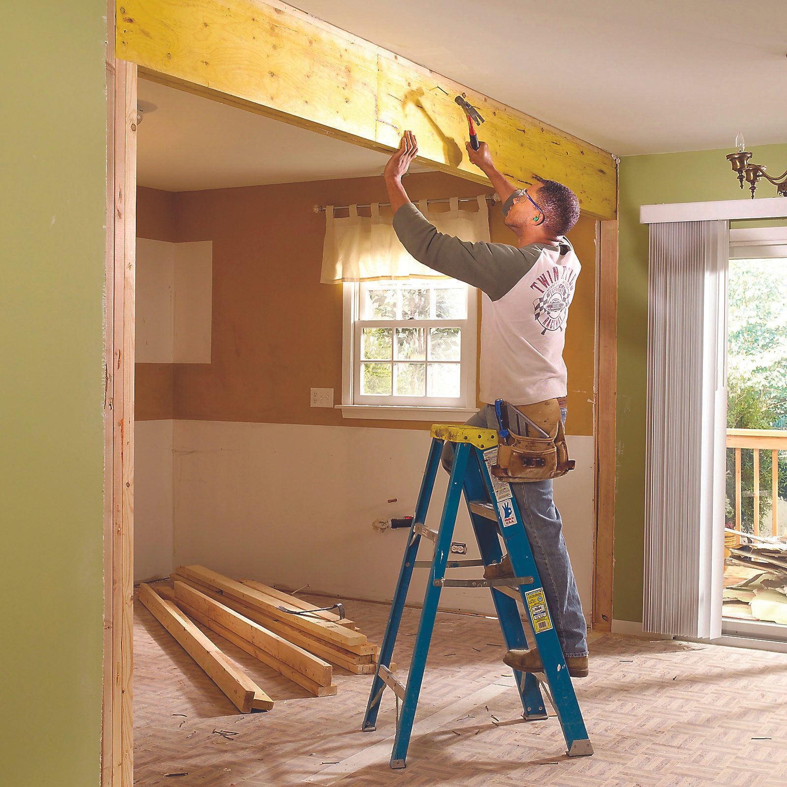 A man is using a hammer to secure a wooden beam while standing on a ladder in a partially renovated room with green walls.