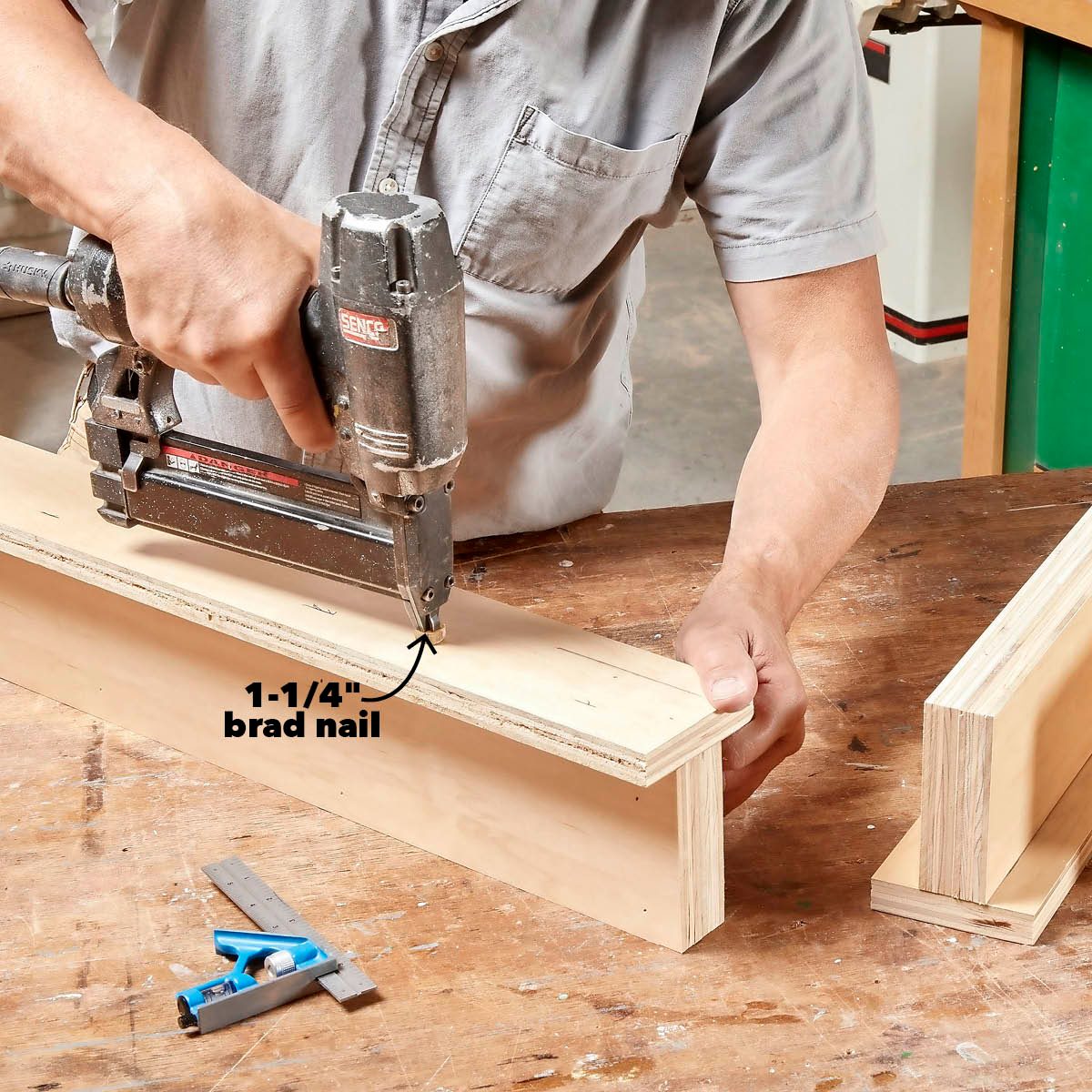 A person uses a nail gun to secure a piece of wood while working at a wooden table, surrounded by tools and another wooden piece.