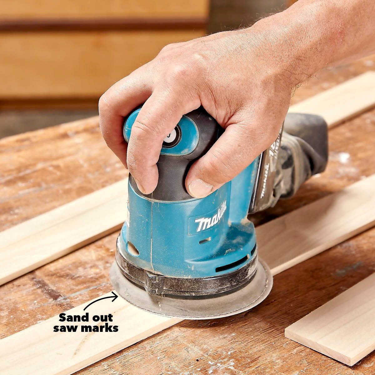 A hand operates a blue power sander on wooden planks, smoothing out imperfections. The workspace is a wooden surface, suggesting a construction or DIY environment.