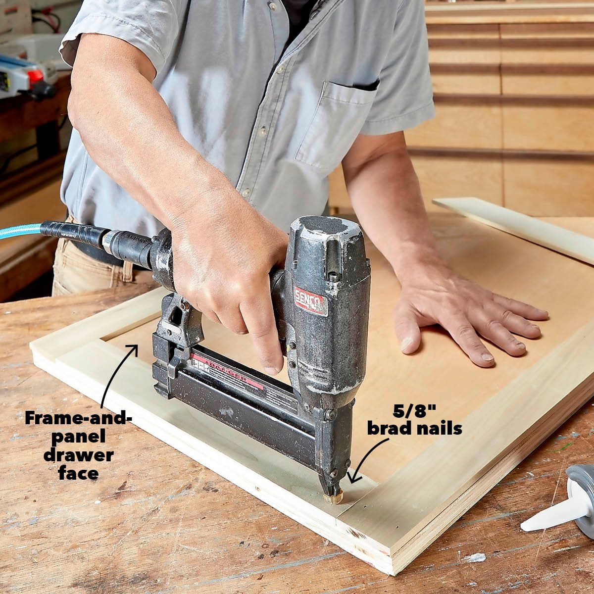 A person uses a brad nailer to attach a frame to a wooden panel on a workbench in a workshop environment.