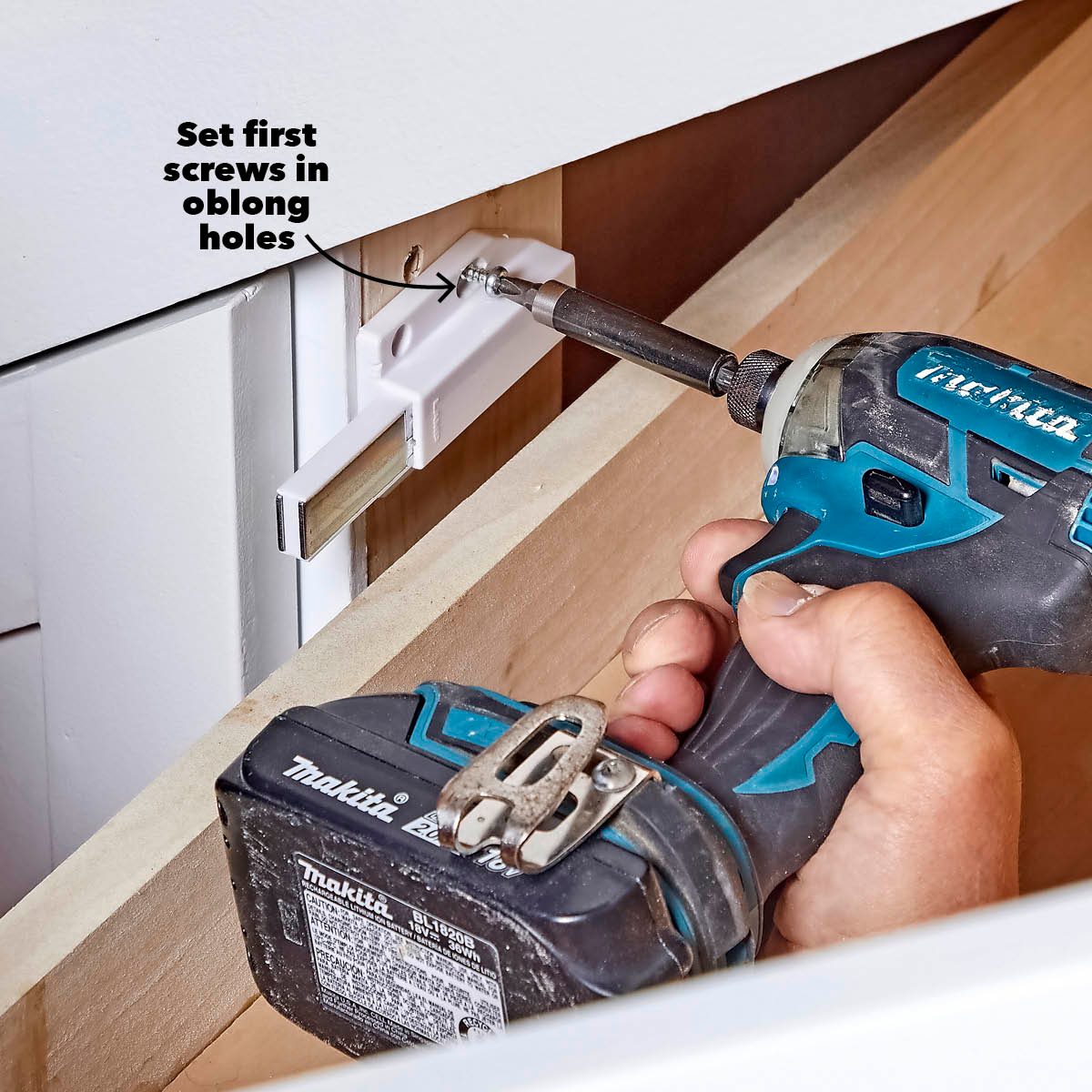 A person uses a drill to secure screws into a mounting bracket inside a wooden drawer, with a visible tool and oblong holes for adjustment.