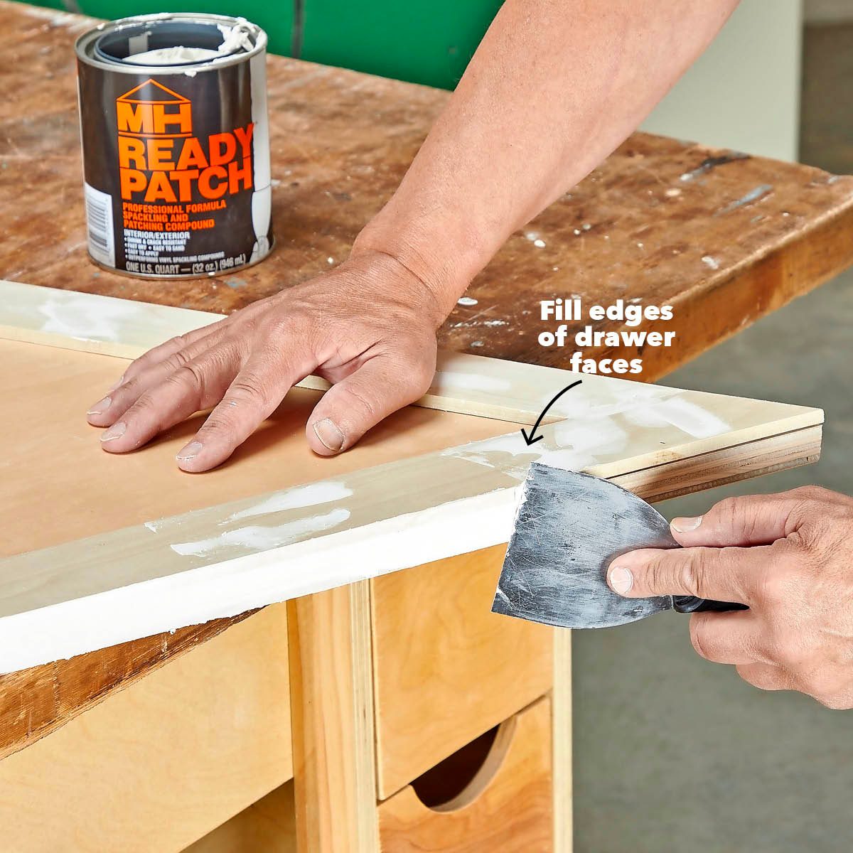 A hand applies a putty knife to fill edges of a drawer face with compound, while another hand holds the surface on a wooden workbench.