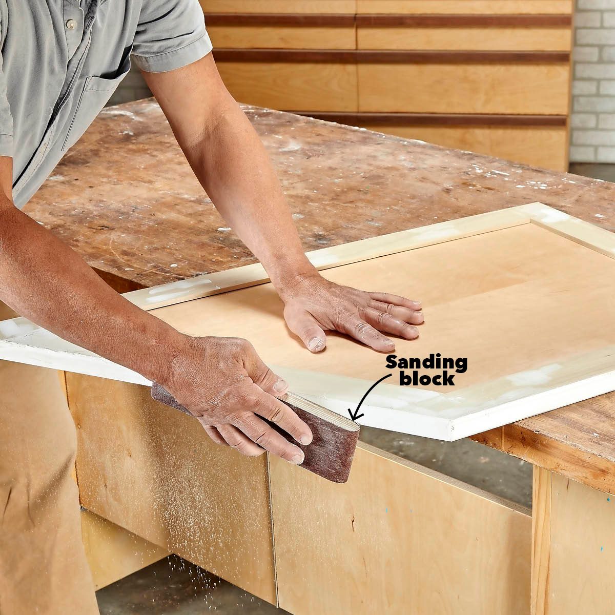 A person's hand uses a sanding block to smooth a wooden frame on a workbench, with wooden cabinets visible in the background.
