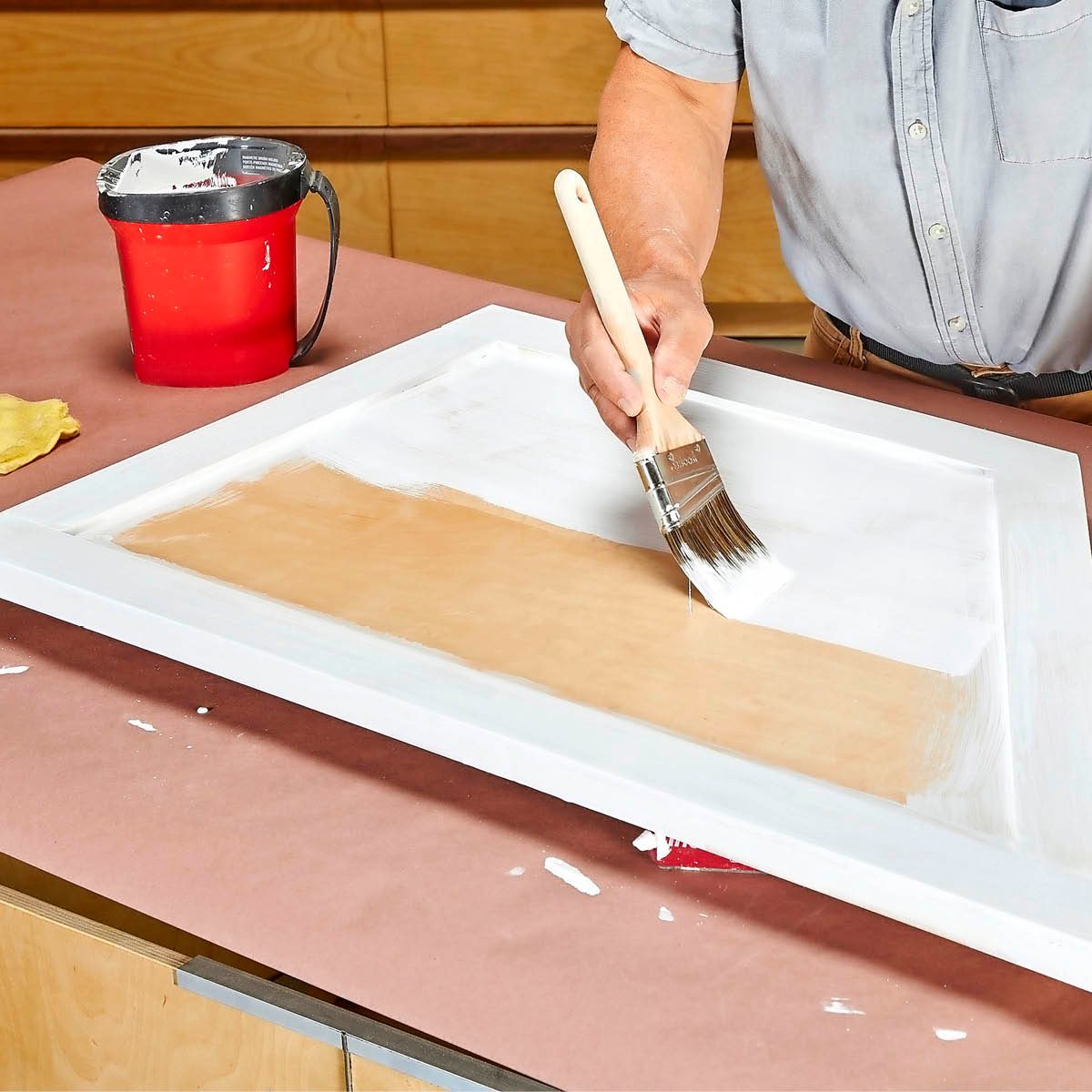 A hand holds a paintbrush, applying white paint to a wooden surface. A red bucket and cloth rest nearby on a brown workspace.