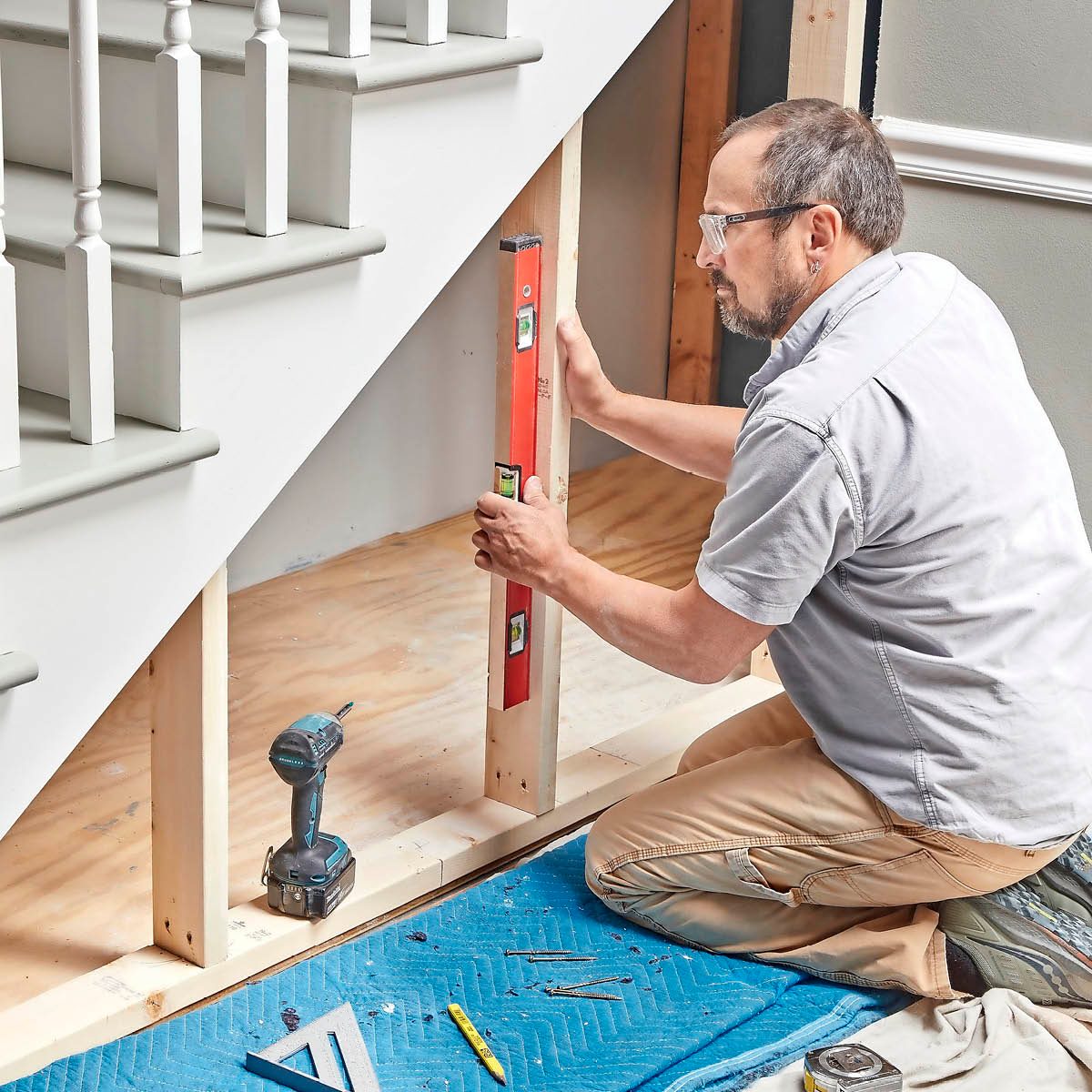 A man uses a level on a wooden frame while kneeling on a blue mat, surrounded by tools and a staircase in a room.