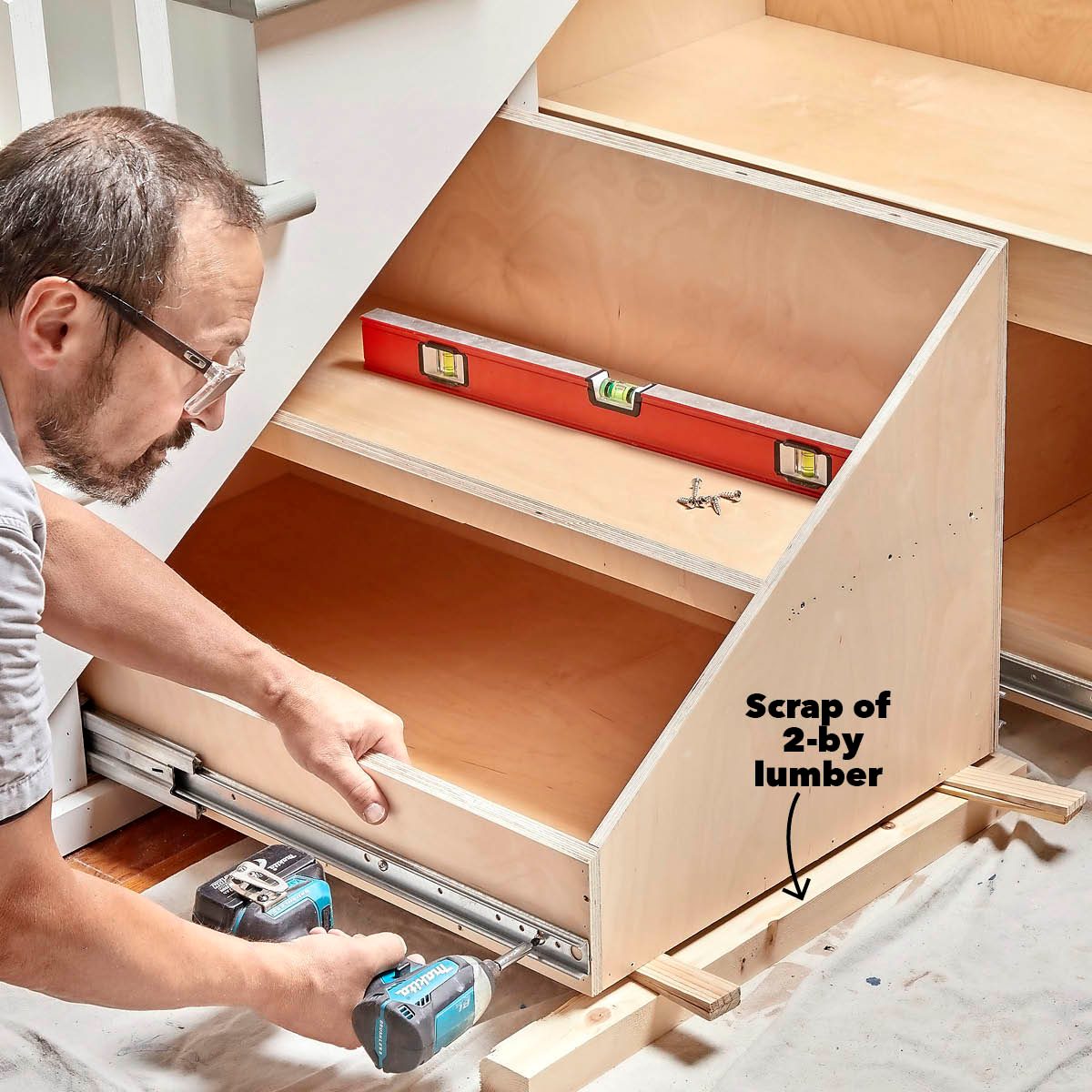 A person installs a drawer slide using a power tool, surrounded by a wooden structure and a level tool, on a protective drop cloth.