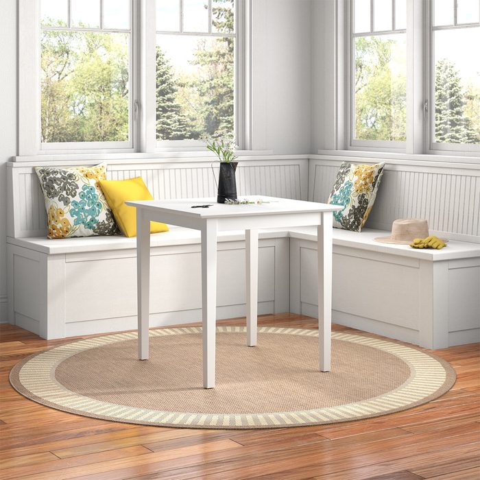 A white table stands in a well-lit corner with cushioned benches, surrounded by large windows and a circular rug, creating a cozy and inviting space.