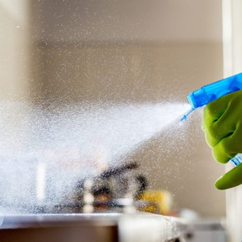 A gloved hand uses a spray bottle, releasing a fine mist of cleaning solution, while a blurred kitchen setting serves as the backdrop.
