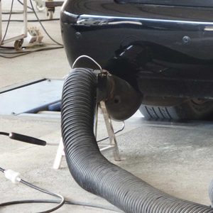 A flexible, coiled vacuum hose connects to the exhaust of a black vehicle, positioned in a garage with concrete flooring and tools in the background.