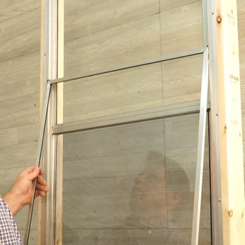 A hand adjusts metal framing for a screen on a wooden wall, reflecting a person's face in the transparent material.