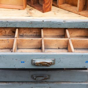 A wooden drawer, partially open, reveals compartments inside. It's positioned on a table, surrounded by other wooden boxes and a vintage setting.