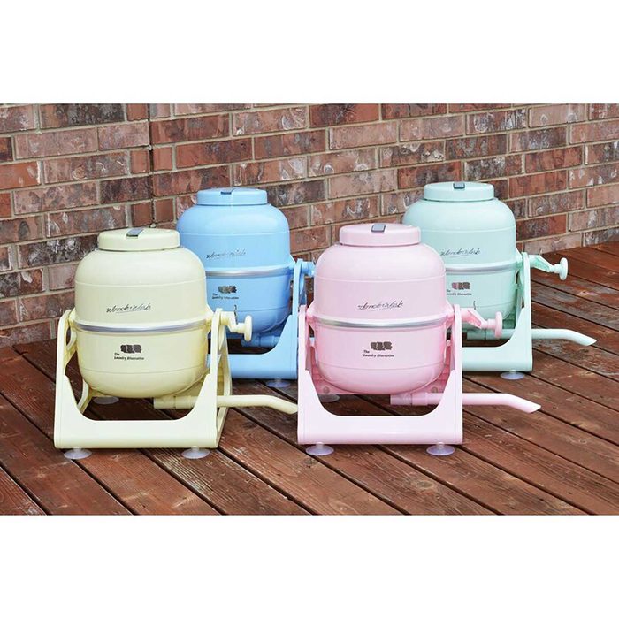 Four pastel-colored laundry wringers are arranged on a wooden deck, each actively positioned for use, set against a backdrop of brick wall.