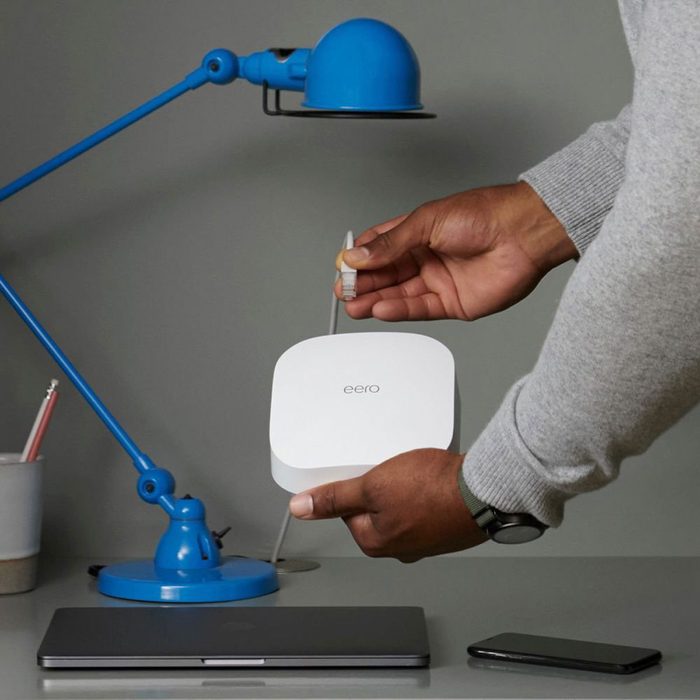 A person connects a cable to a white Eero device on a desk, near a blue lamp, laptop, and smartphone, in a neutral-toned environment.
