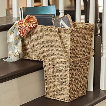 A woven basket containing books and a decorative scarf rests on a staircase, positioned between the steps and a nearby banister.