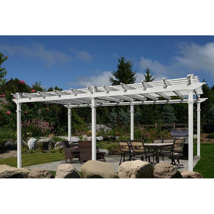 A white pergola stands over a patio with a table and chairs, surrounded by greenery and rocks, under a blue sky with scattered clouds.