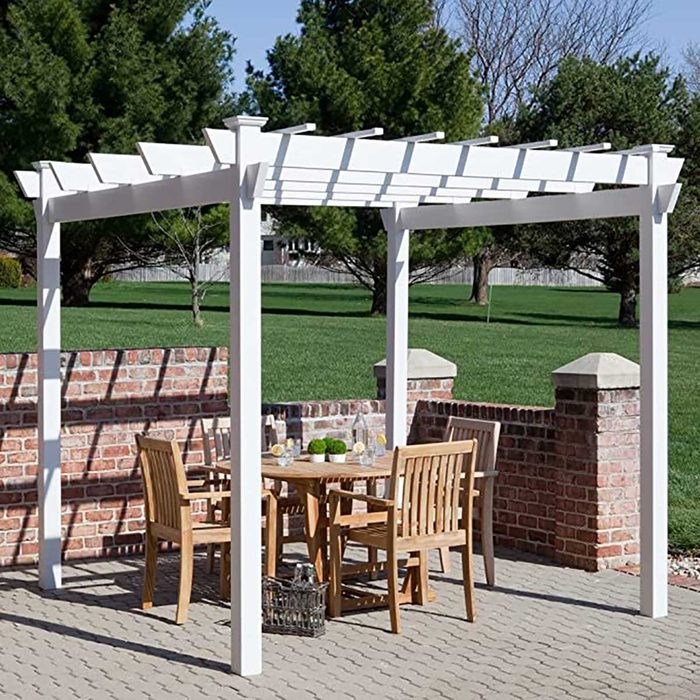 A white pergola shades a wooden table with chairs, set on a stone patio beside a brick wall, surrounded by green grass and trees.