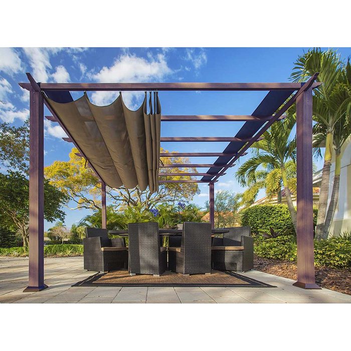 A pergola supports retractable fabric shades above a dark wicker dining set, surrounded by lush greenery and under a blue sky with scattered clouds.