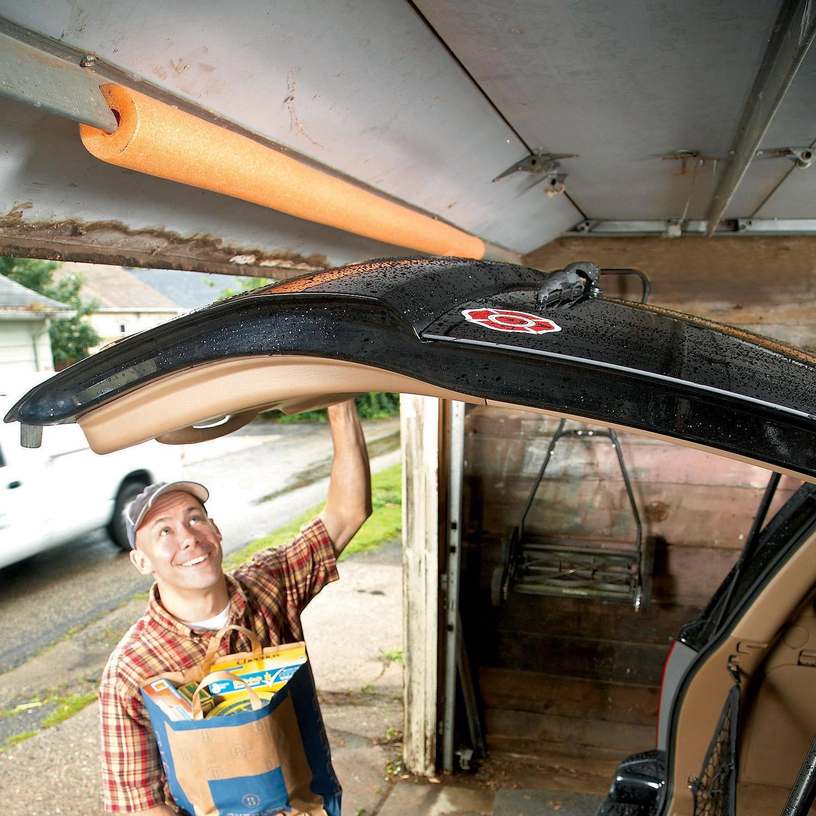 a man with groceries in his hands opening a car hatch door in a garage. The garage door with an orange pool noodle to protect the car