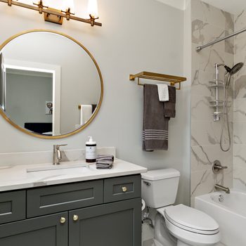 A circular mirror reflects a well-lit bathroom with gray cabinetry, a sink, towels hanging, and a shower setup on marble walls. A bottle sits on the counter.