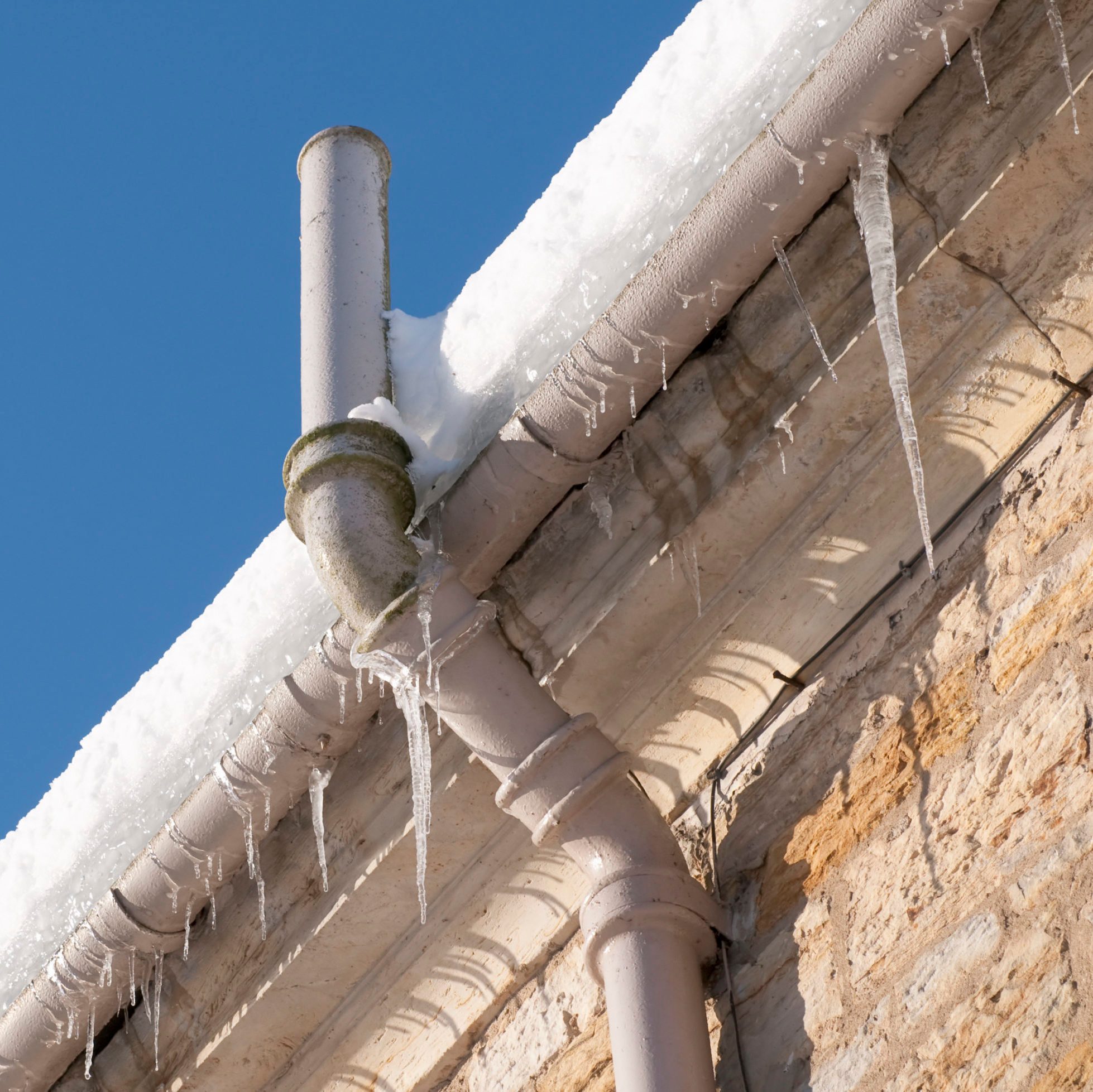 A vertical pipe extends from a snow-covered roof, with icicles hanging underneath, against a clear blue sky and a stone wall backdrop.