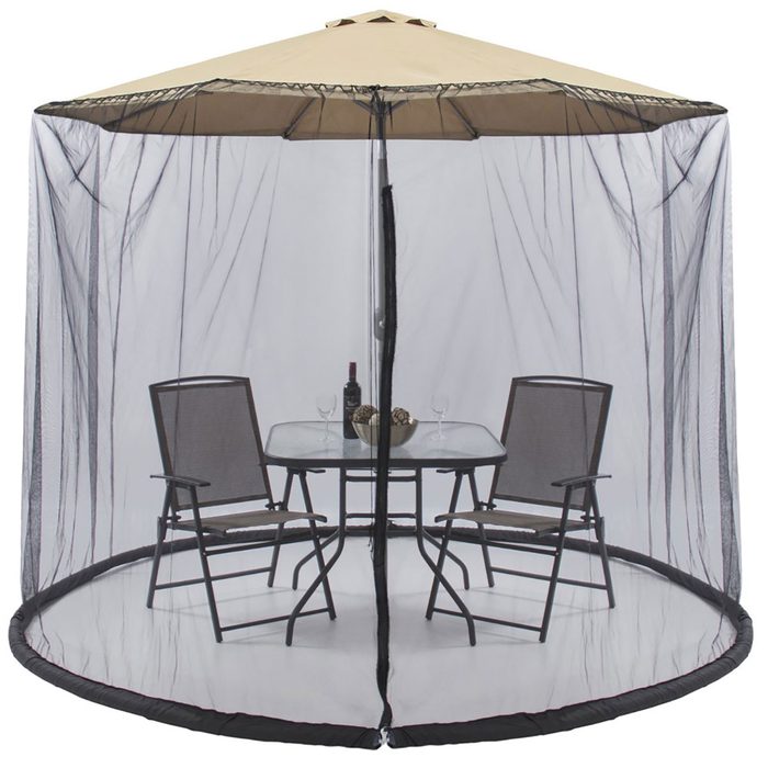 A circular mesh canopy encases a small outdoor dining set with two chairs and a table, shaded by a tan umbrella in an open environment.