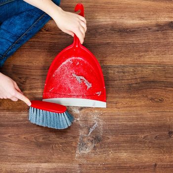A person uses a broom to sweep dirt into a red dustpan while seated on a wooden floor.