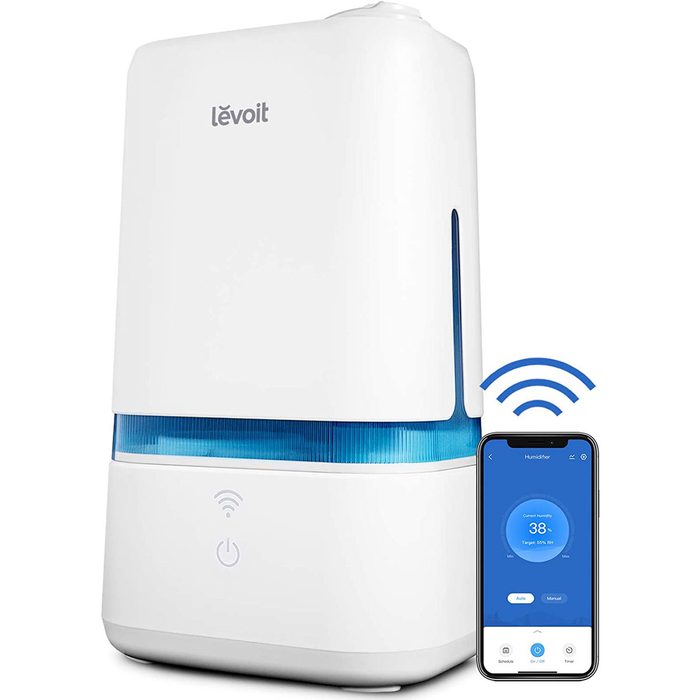 A white humidifier operates, displaying a blue light, while a smartphone app shows humidity levels and control options in a modern indoor setting.