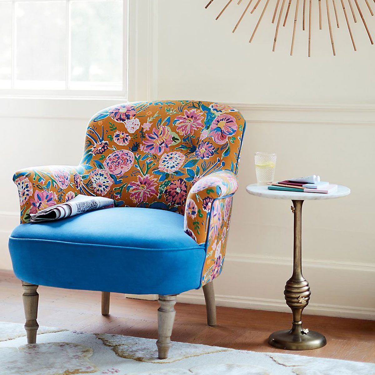 A colorful, upholstered chair with floral patterns sits next to a small, round side table holding a glass and stationery in a bright room.