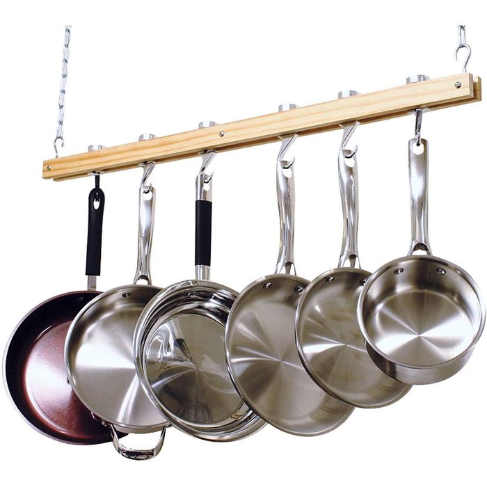 Six hanging frying pans, arranged by size, showcase shiny stainless steel and one copper option, suspended from a wooden rack in a kitchen setting.