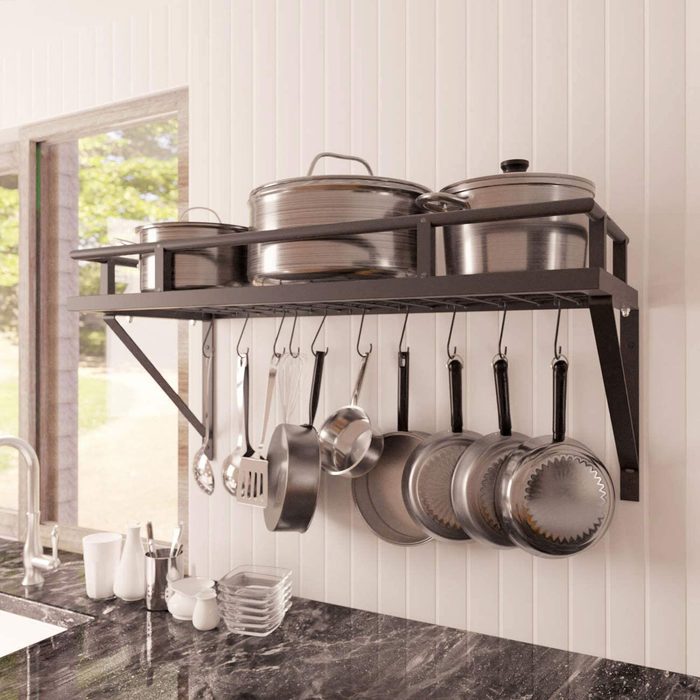 A metal shelf holds several stainless-steel pots above, while various cooking utensils and pans hang below, set against a bright kitchen backdrop.