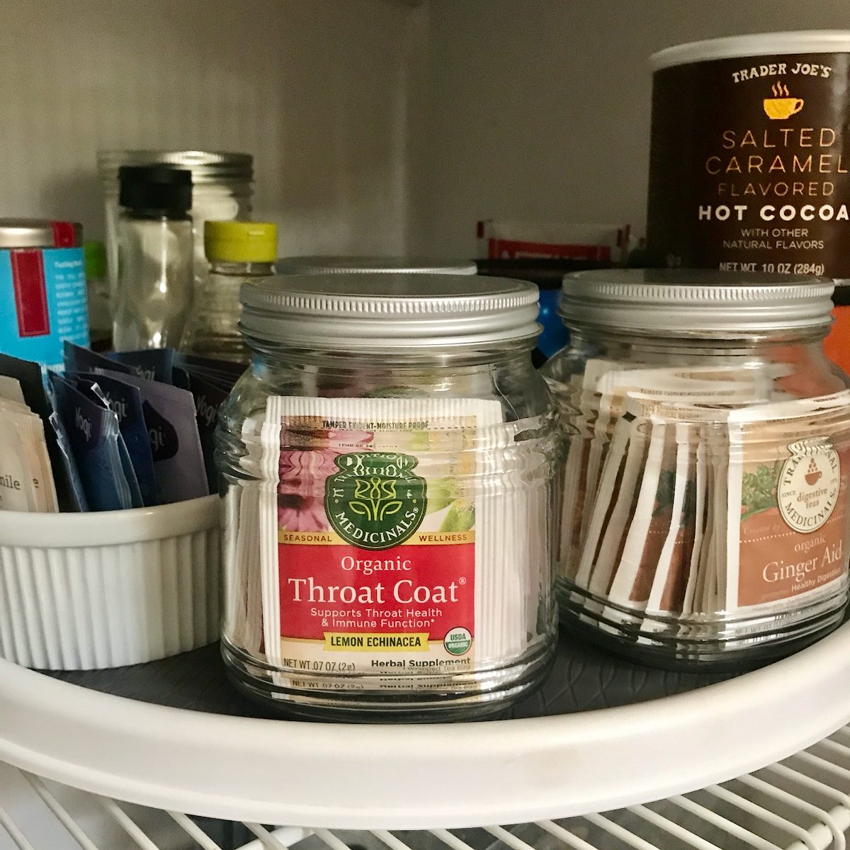 Two glass jars contain herbal tea packets labeled "Organic Throat Coat" and "Ginger Aid," situated on a white turntable in a pantry.