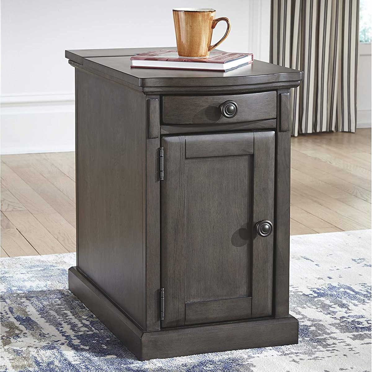 A dark wooden side table stands on a patterned rug, supporting a mug and books, while sunlight filters through nearby striped curtains.