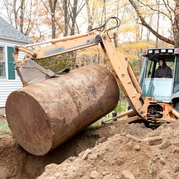 A backhoe excavator lifts a large, rusty tank from a dug-out area beside a house, with autumn trees in the background.