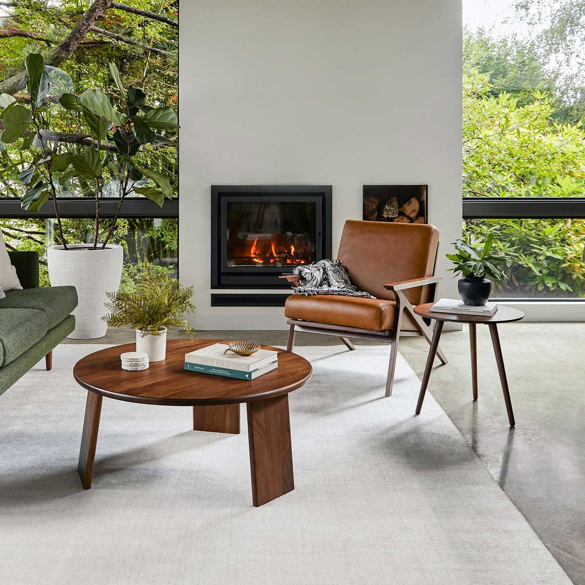 A brown leather chair faces a modern fireplace, with a round wooden coffee table nearby, surrounded by greenery and a spacious, light-filled room.