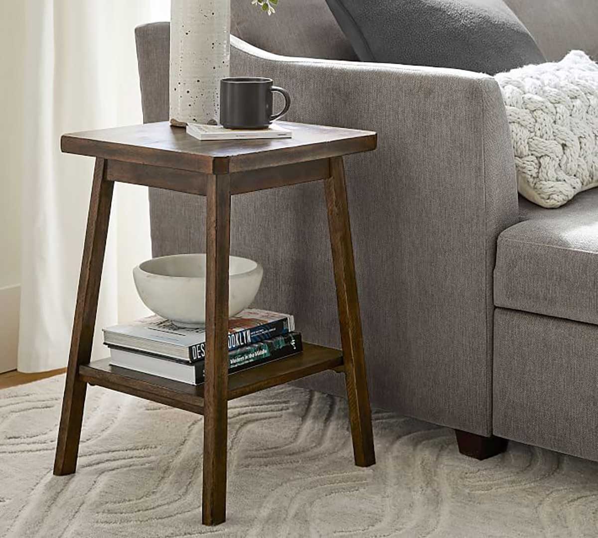 A wooden side table holds a cup, a vase, and books beside a gray couch, on a textured carpet in a softly lit room.