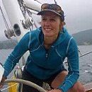A person, smiling, is steering a sailboat on a cloudy day, with water visible in the background, suggesting a maritime adventure.
