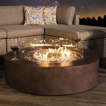A round fire pit with flames burns brightly, surrounded by glass, placed near a cozy sectional sofa and a glass of red wine in an evening setting.