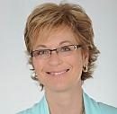 A woman with short, light brown hair and glasses smiles confidently against a neutral background, conveying a sense of approachability and professionalism.