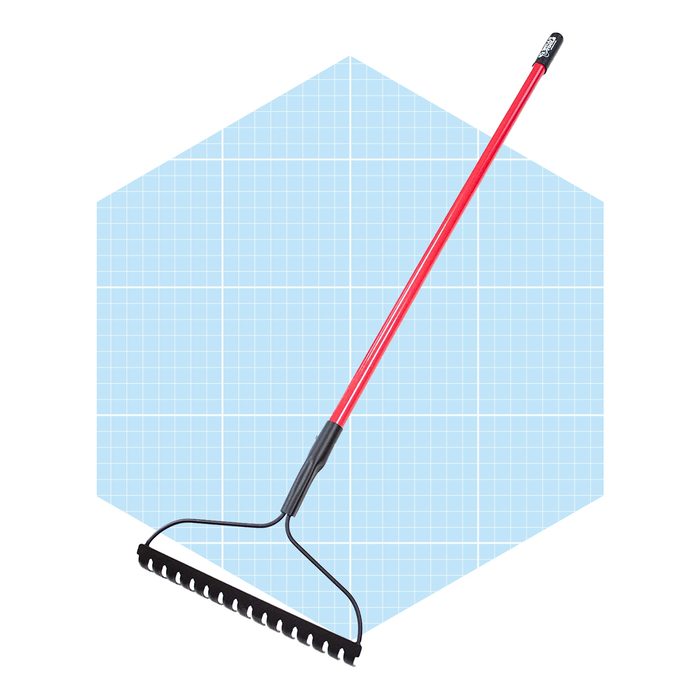A red garden rake rests upright, featuring a black metal head with multiple tines, set against a light blue, grid-patterned background.